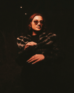 A person with sunglasses and a leopard-print fur jacket stands outdoors at night, dimly lit with hands raised in front of them.